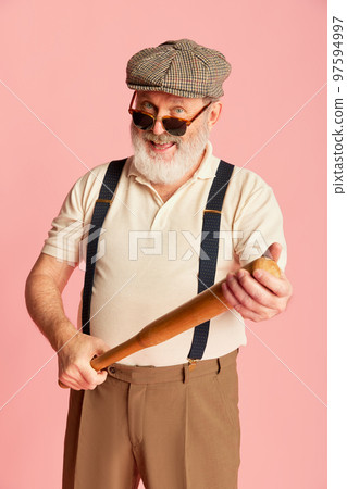 Portrait of senior man in classical clothes, cap, suspenders and sunglasses posing with bat over pink background. Businessman Portrait of senior man in classical clothes, cap, suspenders and sunglasses posing with bat over pink background. Businessman 97594997