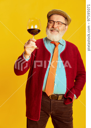 Portrait of senior man in classical clothes, glasses and cap posing with red wine glass over vivid yellow background. Lover of taste Portrait of senior man in classical clothes, glasses and cap posing with red wine glass over vivid yellow background. Lover of taste 97595071
