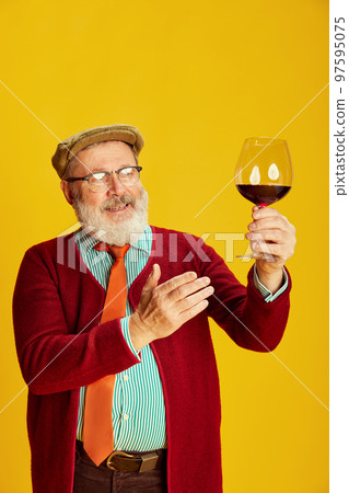 Portrait of senior man in classical clothes, glasses and cap posing, rising glass with red wine over vivid yellow background. Sommelier Portrait of senior man in classical clothes, glasses and cap posing, rising glass with red wine over vivid yellow background. Sommelier 97595075