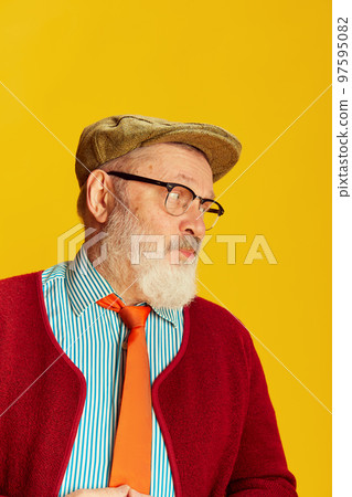 Portrait of senior man in classical clothes, glasses and cap posing over vivid yellow background. Cute look Portrait of senior man in classical clothes, glasses and cap posing over vivid yellow background. Cute look 97595082