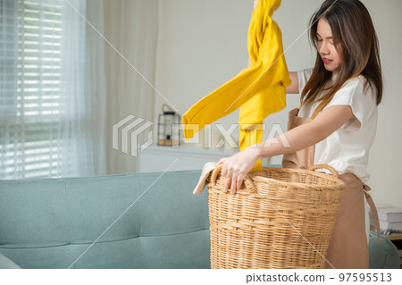 Asian housewife woman holding basket with heap of different clothes on sofa in living room, Beautiful housekeeper doing housework holds wooden basket of clean clothes to washing at home 97595513