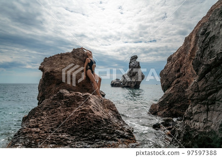 Woman swimsuit sea. Attractive blonde woman in a black swimsuit enjoying the sea air on the seashore around the rocks. Travel and vacation concept. 97597388