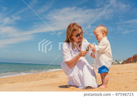 Caucasian mom communicates with a happy one-year-old son on the sea beach in summer. Concept of happy childhood and motherhood. Caucasian mom communicates with a happy one-year-old son on the sea beach in summer. Concept of happy childhood and motherhood. 97598725