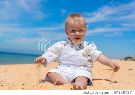 Little happy barefoot boy child sits on the beach and plays with sand on a sunny day. Little happy barefoot boy child sits on the beach and plays with sand on a sunny day. 97598727