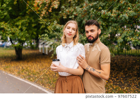 Portrait of happy young couple in love standing holding hands on city park looking at camera. Handsome bearded man with tattoo and cute blonde woman walking drinking coffee enjoying time together. 97607522
