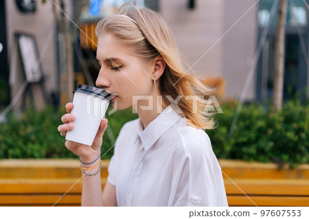 Medium shot of pensive young woman drinking takeaway coffee while waiting someone outdoors in summer day. Elegant blonde lady with closed eyes sitting on bench on urban street with coffee to go. 97607553