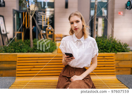 Portrait of confident charming young woman sitting on bench and looking at camera holding mobile phone in hand on background of building. Serious female using mobile phone outdoors in summer. 97607559