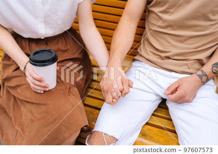 High-angle cropped shot of unrecognizable happy young couple in love holding hands sitting on bench and drinking takeaway coffee enjoying time together, outdoors on summer day. 97607573