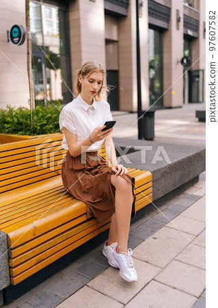 Vertical shot of serious young blonde woman wearing casual clothes sitting on yellow bench alone and checking notifications on mobile phone, on background of building on street in summer day. 97607582