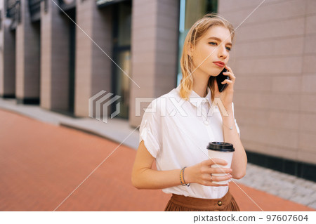 Medium shot of elegant blonde female teenager in casual clothes holding takeaway coffee cup and talking on smartphone during walking on city street in sunny summer day, looking away. 97607604