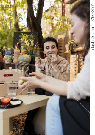 Happy young woman customer enjoying tasting organic fruits and veggies at local food market on sunny autumn day. Female farmer sitting at table with consumer, offering to taste homegrown produce 97608457
