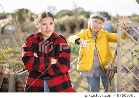 Two angry casual women neighbors of different ages arguing during the vegetable garden season on sunny day of spring Two angry casual women neighbors of different ages arguing during the vegetable garden season on sunny day of spring 97609503