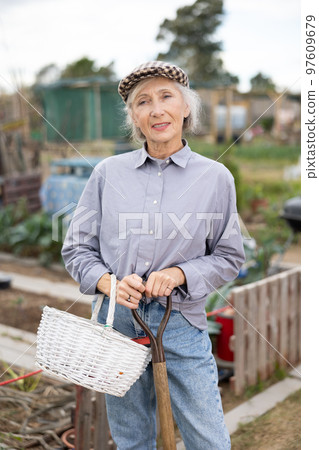 Happy mature female holding wicker basket and handle of shovel while standing in smallholding 97609679