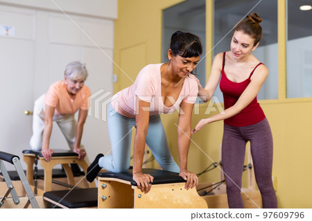 Young woman pilates instructor helping Hispanic woman exercising on combo chair Young woman pilates instructor helping Hispanic woman exercising on combo chair 97609796