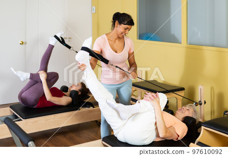 Active women perform Pilates exercises on a simulator with the help of a trainer Active women perform Pilates exercises on a simulator with the help of a trainer 97610092