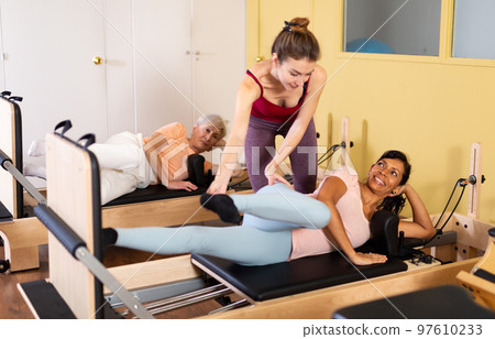 Young pilates instructor assisting Latin American woman exercising on reformer Young pilates instructor assisting Latin American woman exercising on reformer 97610233
