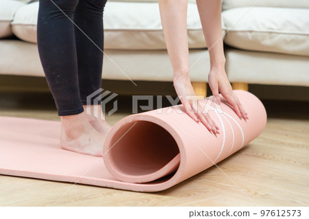 Close-up of attractive young woman folding pink yoga or fitness mat after working out at home in living room. 97612573