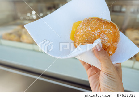 Hand holding Bola de Berlim or Berlim Ball, a Portuguese pastry made from a fried donut filled with sweet eggy cream and rolled in crunchy sugar. Hand holding Bola de Berlim or Berlim Ball, a Portuguese pastry made from a fried donut filled with sweet eggy cream and rolled in crunchy sugar. 97618325