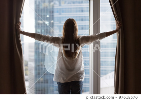 Rear view of young homeowner woman opening drapes at window 97618570