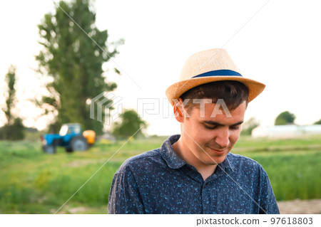 Defocus farmer in hat working on field with greenhouse outdoor. Portrait of young Caucasian handsome happy man farmer standing in field and smiling. Tractor on background. Out of focus Defocus farmer in hat working on field with greenhouse outdoor. Portrait of young Caucasian handsome happy man farmer standing in field and smiling. Tractor on background. Out of focus 97618803