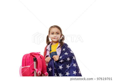 Charming little girl, a smart school kid, wrapping in USA flag, standing near a backpack, looking at camera, on white background. Concept of studying abroad. English courses. British American schools Charming little girl, a smart school kid, wrapping in USA flag, standing near a backpack, looking at camera, on white background. Concept of studying abroad. English courses. British American schools 97619701