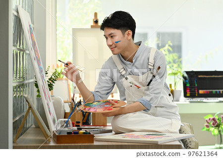Happy asian male artist holding paintbrushes and palette painting on canvas at art studio. Education, hobby, art concept Happy asian male artist holding paintbrushes and palette painting on canvas at art studio. Education, hobby, art concept 97621364