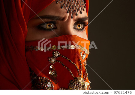 Studio shot of a young charming woman wearing the terracotta hijab decorated with sequins and jewelry. Arabic style. 97621476