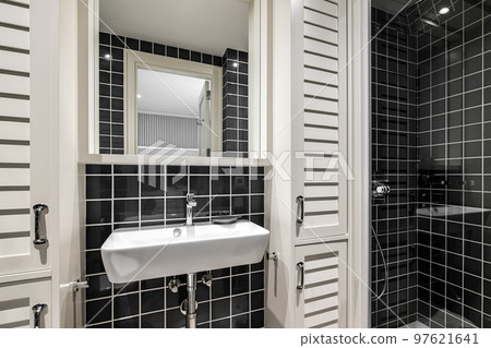 Trendy black ceramic tile bathroom creates striking contrast with white sink and light wood lockers. Mirror reflects entrance to room. Shower area is illuminated by ceiling lights. Trendy black ceramic tile bathroom creates striking contrast with white sink and light wood lockers. Mirror reflects entrance to room. Shower area is illuminated by ceiling lights. 97621641