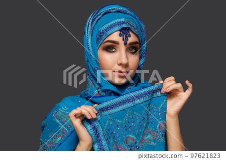 Studio shot of a young beautiful woman wearing the blue hijab decorated with sequins and jewelry. Arabic style. 97621823