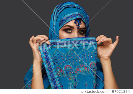Studio shot of a young beautiful woman wearing the blue hijab decorated with sequins and jewelry. Arabic style. 97621824