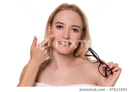 Young blonde woman holding contact lens on finger in front of her face and holding in her other hand a black glasses on white background. Young blonde woman holding contact lens on finger in front of her face and holding in her other hand a black glasses on white background. 97621893