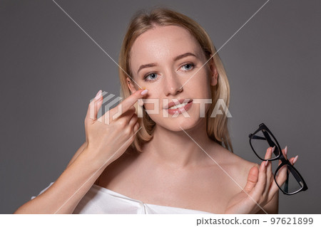 Young blonde woman holding contact lens on finger in front of her face and holding in her other hand a black glasses on gray background. 97621899