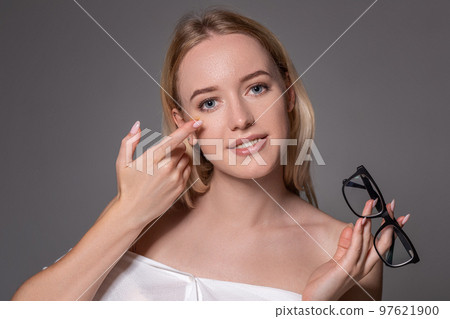 Young blonde woman holding contact lens on finger in front of her face and holding in her other hand a black glasses on gray background. Young blonde woman holding contact lens on finger in front of her face and holding in her other hand a black glasses on gray background. 97621900