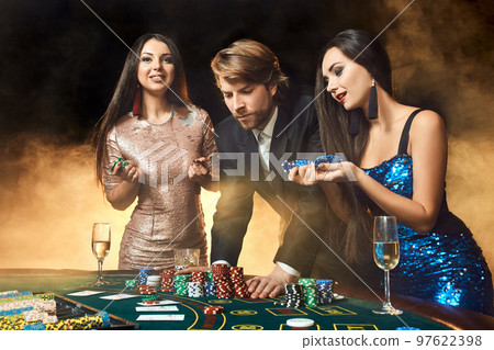 Two beautiful women and young man play on poker table in casino, focus on man and brunette 97622398