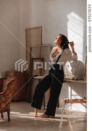 a brunette in a white shirt and trousers with suspenders in the artist's studio. 97623634