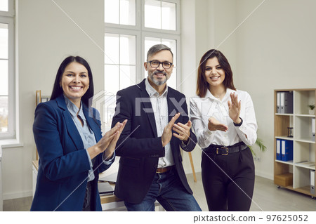 Brutal, gray-haired, happy man and two beautiful, smiling young businesswomen applaud looking at the camera. 97625052