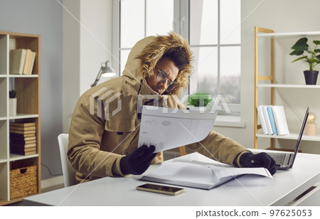 Man in a winter clothes looking at heating bills while sitting at his desk in his cold house Man in a winter clothes looking at heating bills while sitting at his desk in his cold house 97625053