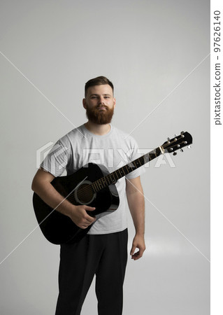 Male musician playing on acoustic guitar. Portraite of bearded handsome musician holding a guitar in a white room. Guitarist, artist, composer, rocker, star. Male musician playing on acoustic guitar. Portraite of bearded handsome musician holding a guitar in a white room. Guitarist, artist, composer, rocker, star. 97626140