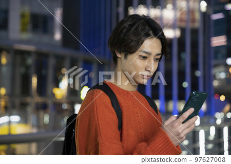 A young man looking at a smartphone screen on a street corner at night 97627008