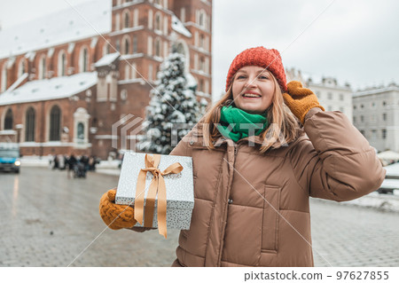 Happy blonde woman smiling confident surprise with Christmas gift outdoors at Christmas market on town hall square in Krakow, Poland. The girl looks at the decorated Christmas tree in the city square 97627855