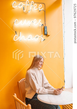 Attractive young blonde smiling woman with cup of coffee using mobile phone in cafe on a yellow background Attractive young blonde smiling woman with cup of coffee using mobile phone in cafe on a yellow background 97629725