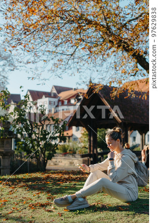 Young Fashionable Teenage Girl With Smartphone In Europian Park In Autumn Sitting At Smiling. Trendy Young Woman In Fall In Park Texting. Retouched, Vibrant Colors. Beautiful Blonde Teenage Girl 97629838