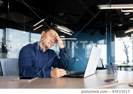 A young Asian man, an office worker, feels a strong headache. Migraine. He sits at the desk, holds his head, closed his eyes, took off his glasses, and rests. A young Asian man, an office worker, feels a strong headache. Migraine. He sits at the desk, holds his head, closed his eyes, took off his glasses, and rests. 97631017