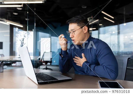 Young male Asian office worker having asthma attack at workplace, breathing hard. He sits at a table with a laptop, uses an inhaler. Young male Asian office worker having asthma attack at workplace, breathing hard. He sits at a table with a laptop, uses an inhaler. 97631018