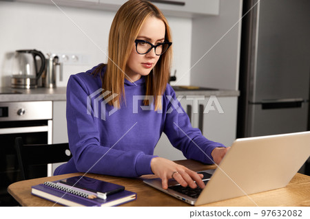 Pensive young adult woman in glasses work study at kitchen using laptop. Remote education 97632082