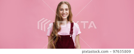 Waist-up shot of kind friendly-looking pleasant young female student with long wavy fair hair in trendy overalls wearing makeup smiling joyfully and gazing at camera with good mood over pink wall Waist-up shot of kind friendly-looking pleasant young female student with long wavy fair hair in trendy overalls wearing makeup smiling joyfully and gazing at camera with good mood over pink wall 97635809