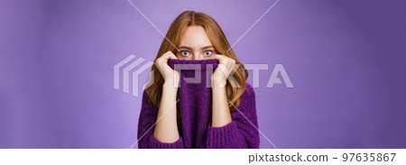 Redhead girl scared winter is comming hiding in collar of warm sweater popping eyes shocked and stunned at camera as if frightened and insecure standing terrified over violet background Redhead girl scared winter is comming hiding in collar of warm sweater popping eyes shocked and stunned at camera as if frightened and insecure standing terrified over violet background 97635867