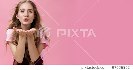 Portrait of sensual and tender gentle young wman with wavy natural hair bending towards camera facing wind holding palms near folded lips sending air kiss forward with flirty caring and loving look 97636592