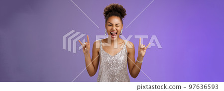 Excited good-looking african-american woman in glittering silver shiny dress having fun amusing university party prom night show tongue winking joyfully rock-n-roll heavy-metal gesture, blue wall 97636593
