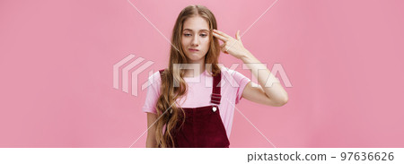 Portrait of bothered uneasy cute european female with long natural fair hair in trendy corduroy overalls smirking from irritation and annoyance showing finger fun gesture as if shooting in head Portrait of bothered uneasy cute european female with long natural fair hair in trendy corduroy overalls smirking from irritation and annoyance showing finger fun gesture as if shooting in head 97636626